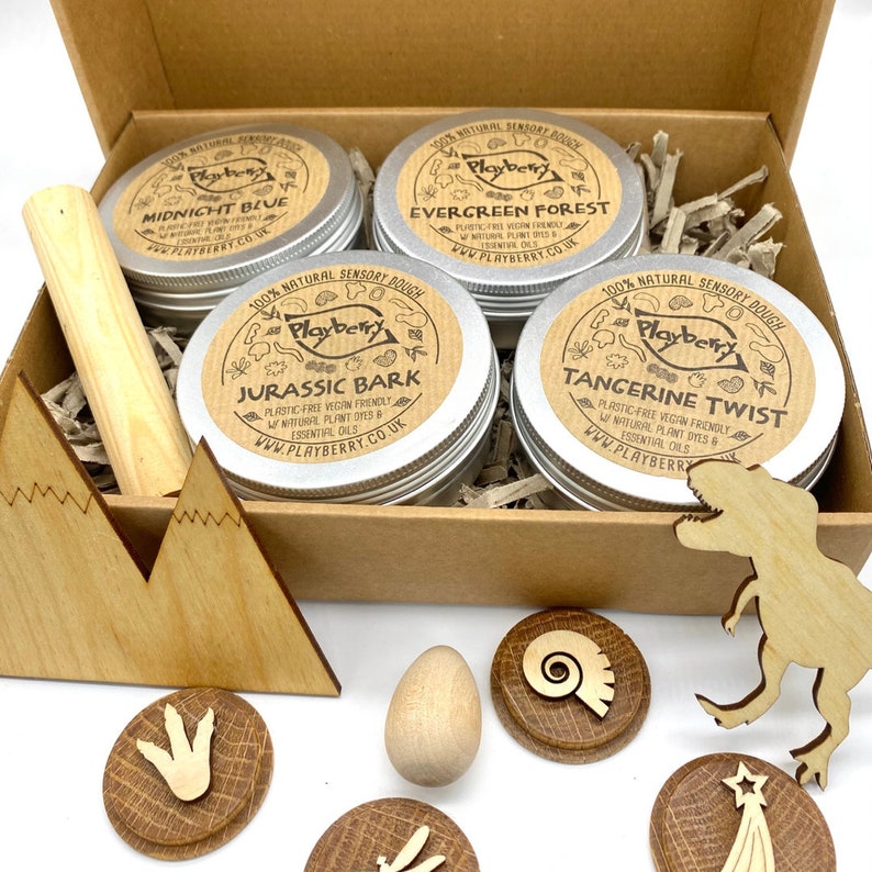 K&ouml;nnte beinhalten: Ein Set mit vier kleinen, silbernen Dosen mit nat&uuml;rlichem sensorischem Teig, beschriftet mit "Midnight Blue", "Evergreen Forest", "Jurassic Bark" und "Tangerine Twist". Die Dosen befinden sich in einer braunen Pappbox mit Holz-Dinosaurierspielzeug und einem Holzei.