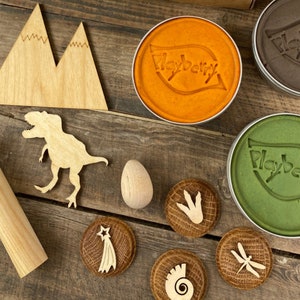 K&ouml;nnte beinhalten: Ein Set aus Holzspielzeug f&uuml;r Knetmasse zum Thema Dinosaurier, bestehend aus einem Dinosaurier, einem Berg, einem Fu&szlig;abdruck, einer Muschel, einem Sternschnuppen, einer Libelle und einem Ei. Die Knetmasse befindet sich in orangefarbenen, gr&uuml;nen und braunen Beh&auml;ltern mit dem Playberry-Logo.