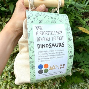 K&ouml;nnte beinhalten: Eine gr&uuml;n-wei&szlig;e Tasche mit einem Holzgriff. Die Tasche hat ein Etikett mit der Aufschrift "A Storyteller's Sensory Talekit Dinosaurs". Auf dem Etikett steht auch "Creative play made from plants! Eco-friendly sensory dough kit with themed wooden accessories to encourage big imaginations for little hands."