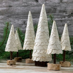 May include: A collection of six decorative paper Christmas trees. The trees are cone-shaped, made from pages of a book, and mounted on small wooden bases. The trees vary in size and are arranged on a wooden surface.