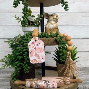 May include: A three-tiered wooden tiered tray with a black metal stand. The top tier has a bird's nest with a blue egg, a small potted plant with a "bloom" sign, and a small rabbit figurine. The middle tier has a floral tag, a wooden bead garland, and a potted succulent. The bottom tier has a rolling pin wrapped in floral fabric and a potted succulent.