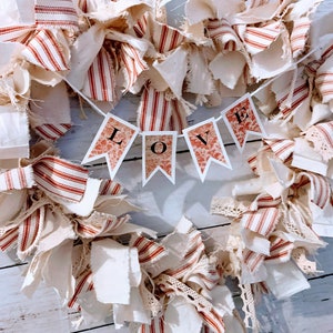 May include: A heart-shaped wreath made of white and red striped fabric, with a banner that reads "LOVE" in the center. The wreath is decorated with white fabric scraps and lace.