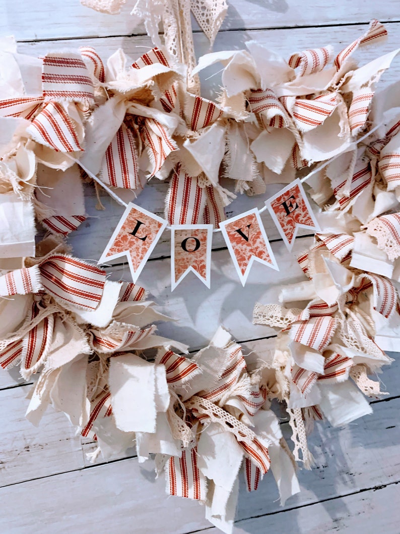 May include: A heart-shaped wreath made of white and red striped fabric, with a banner that reads "LOVE" in the center. The wreath is decorated with white fabric scraps and lace.