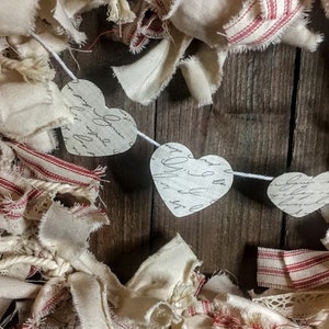 May include: A white fabric garland with red stripes and lace trim. The garland is decorated with paper hearts that have handwritten script on them. The garland is perfect for Valentine's Day or a rustic wedding.