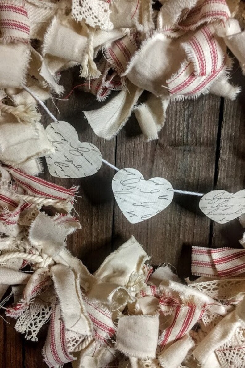 May include: A white fabric garland with red stripes and lace trim. The garland is decorated with paper hearts that have handwritten script on them. The garland is perfect for Valentine's Day or a rustic wedding.