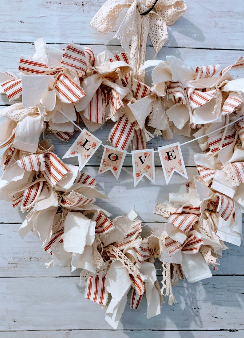 May include: A heart-shaped wreath made of white and red striped fabric, white fabric, and lace. The wreath has a banner that says "LOVE" in black letters.