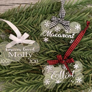 May include: Three clear acrylic dog bone ornaments with white snowflake designs and white lettering. The ornaments are decorated with red and black checked ribbon and white ribbon. The ornaments are personalised with the names "Macaroni", "Molly", and "Elliot" and the dates "2002" and "2013".