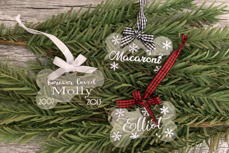 May include: Three clear acrylic dog bone ornaments with white snowflake designs and white lettering. The ornaments are decorated with red and black checked ribbon and white ribbon. The ornaments are personalised with the names "Macaroni", "Molly", and "Elliot" and the dates "2002" and "2013".