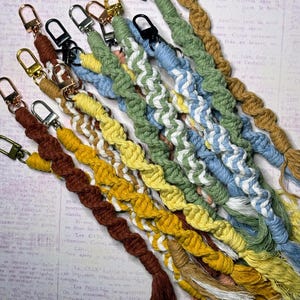 May include: A selection of macrame keychains in shades of brown, yellow, green, and blue. Each keychain has a braided design and a metal clasp for attaching keys or accessories. The keychains are handmade.