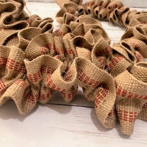 May include: A close-up of a rustic burlap wreath with red accents. The wreath is made of woven burlap fabric, with a ruffled design. The red accents are in a repeating pattern. The wreath is a decorative item.