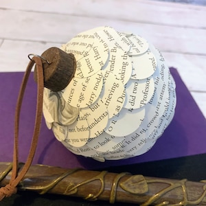 May include: A brown and gold wand with a leafy design. The wand is lying on a purple surface next to a white paper ornament decorated with text from a book. The ornament is hanging from a brown leather cord.