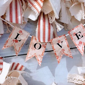 May include: A white string banner with red and white fabric scraps in the background. The banner has four flags with the word "LOVE" printed in black letters.