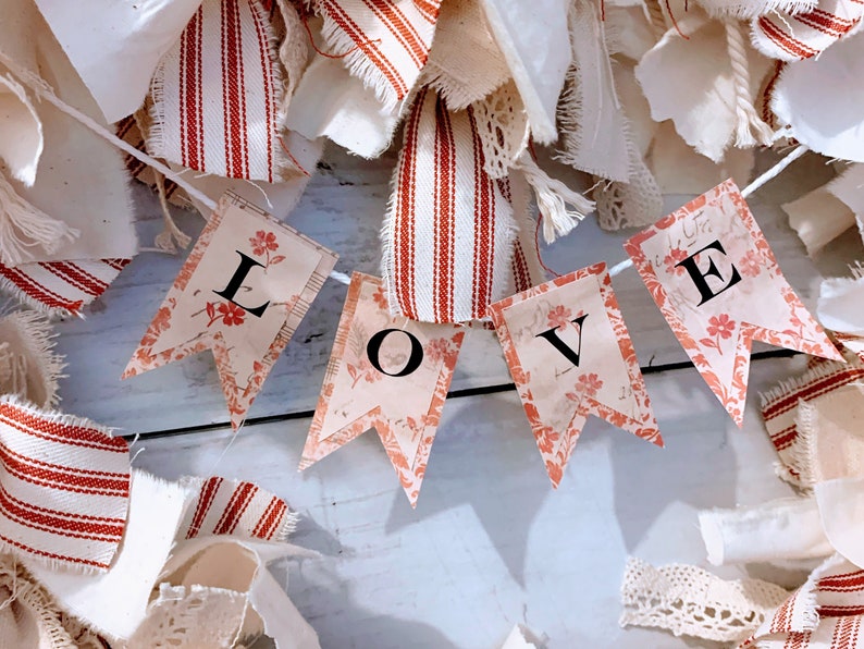 May include: A white string banner with red and white fabric scraps in the background. The banner has four flags with the word "LOVE" printed in black letters.