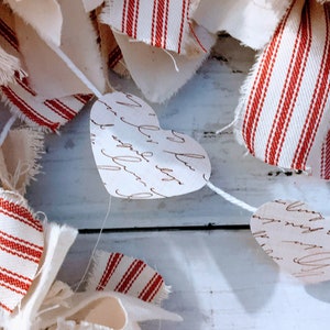 May include: A white string garland with red and white striped fabric and paper hearts with handwritten script. The garland is hanging on a white wooden surface.
