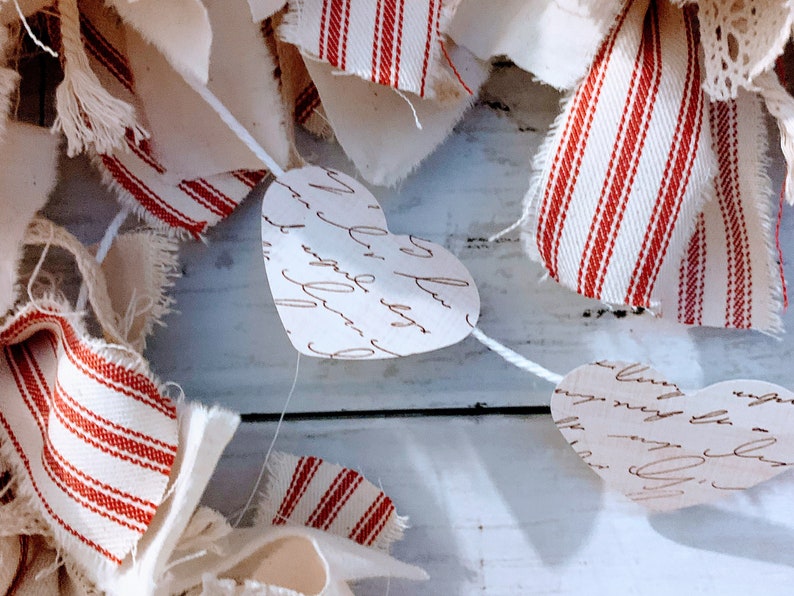 May include: A white string garland with red and white striped fabric and paper hearts with handwritten script. The garland is hanging on a white wooden surface.