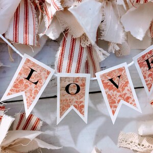 May include: A white string banner with the word "LOVE" printed on it in black letters. The banner is made of paper and has a red and white floral pattern. The banner is hanging from a white string and is surrounded by white and red fabric scraps.