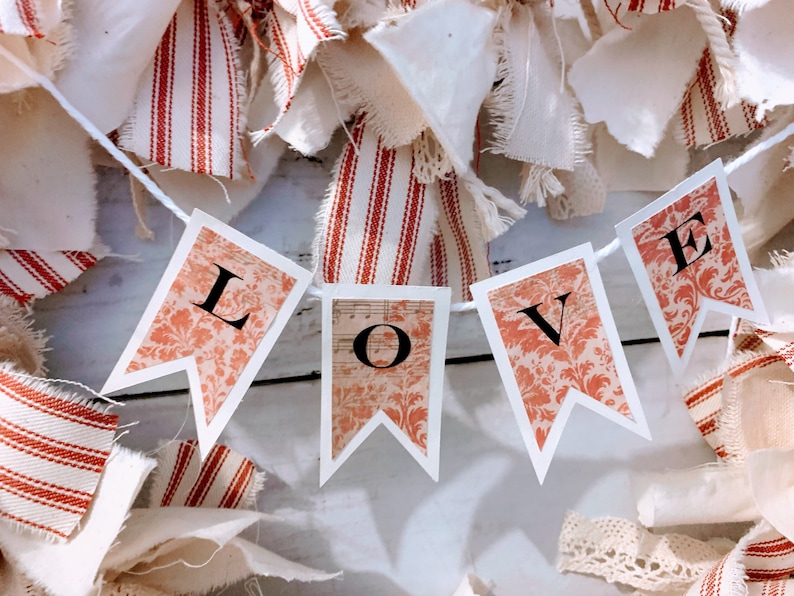 May include: A white string banner with the word "LOVE" printed on it in black letters. The banner is made of paper and has a red and white floral pattern. The banner is hanging from a white string and is surrounded by white and red fabric scraps.