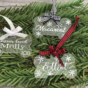 May include: Three clear acrylic bone-shaped Christmas decorations with white snowflakes and personalised text. The decorations are adorned with black and white checked ribbon, red and white checked ribbon, and a white ribbon. The text on the decorations reads "Macaroni", "Elliot", and "forever loved Molly 2002 - 2013".