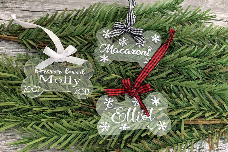 May include: Three clear acrylic bone-shaped Christmas decorations with white snowflakes and personalised text. The decorations are adorned with black and white checked ribbon, red and white checked ribbon, and a white ribbon. The text on the decorations reads "Macaroni", "Elliot", and "forever loved Molly 2002 - 2013".