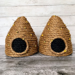 May include: Two handmade, cone-shaped pet houses made of woven brown rope. Each house has a circular black opening and is adorned with small, yellow and black bee decorations. The houses are designed to resemble beehives.