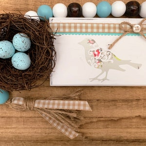 May include: A white wooden sign with a blue and white checkered ribbon and a brown twine bow. The sign features a white silhouette of a bird with floral accents and the word "TWEET" on a blue and white wooden tag. A nest with three blue speckled eggs sits on a wooden surface with a blue and white beaded garland.