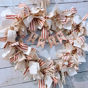 May include: A heart-shaped wreath made of white and red striped fabric, white fabric, and lace. The wreath has a banner that says "LOVE" in black letters.