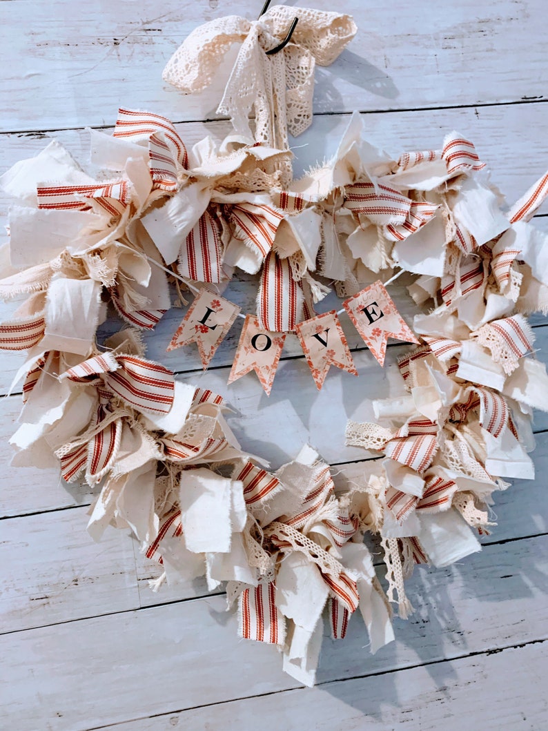 May include: A heart-shaped wreath made of white and red striped fabric, white fabric, and lace. The wreath has a banner that says "LOVE" in black letters.