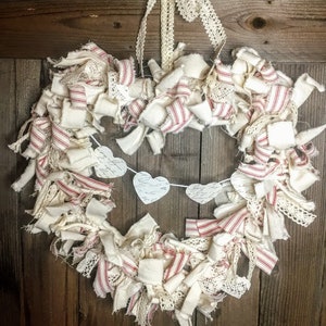 May include: A heart-shaped wreath made of white fabric strips with red and white stripes and white lace. The wreath is decorated with a banner of white paper hearts with handwritten calligraphy.