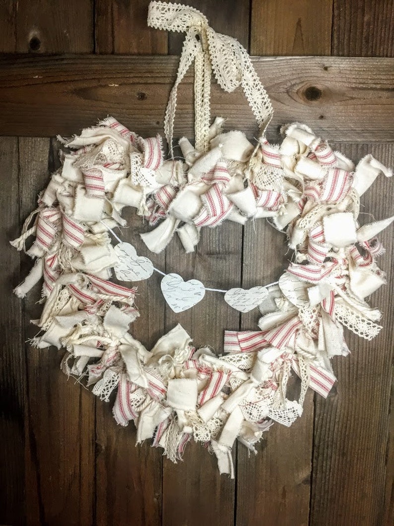May include: A heart-shaped wreath made of white fabric strips with red and white stripes and white lace. The wreath is decorated with a banner of white paper hearts with handwritten calligraphy.