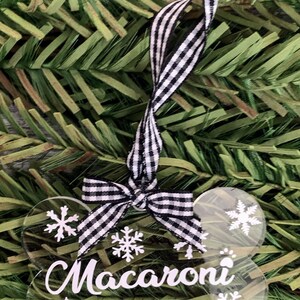 May include: A clear plastic bone-shaped Christmas bauble with a black and white checked ribbon. The bauble has the name "Macaroni" written on it and is decorated with white snowflakes.