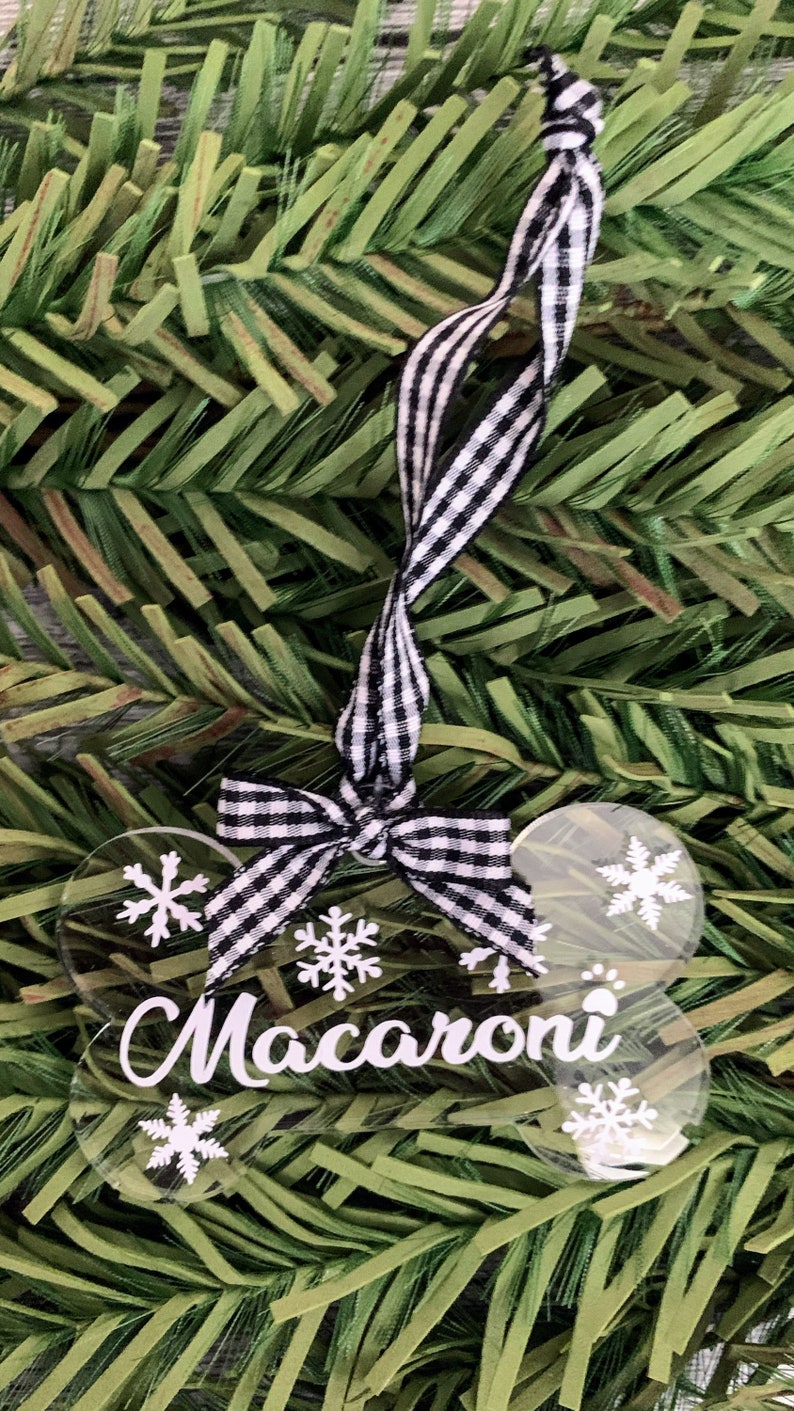 May include: A clear plastic bone-shaped Christmas bauble with a black and white checked ribbon. The bauble has the name "Macaroni" written on it and is decorated with white snowflakes.