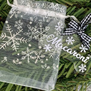 May include: A clear acrylic bone-shaped Christmas bauble with the name "Macaroni" written on it in white. The bauble is decorated with white snowflakes and is tied with a black and white checked ribbon.