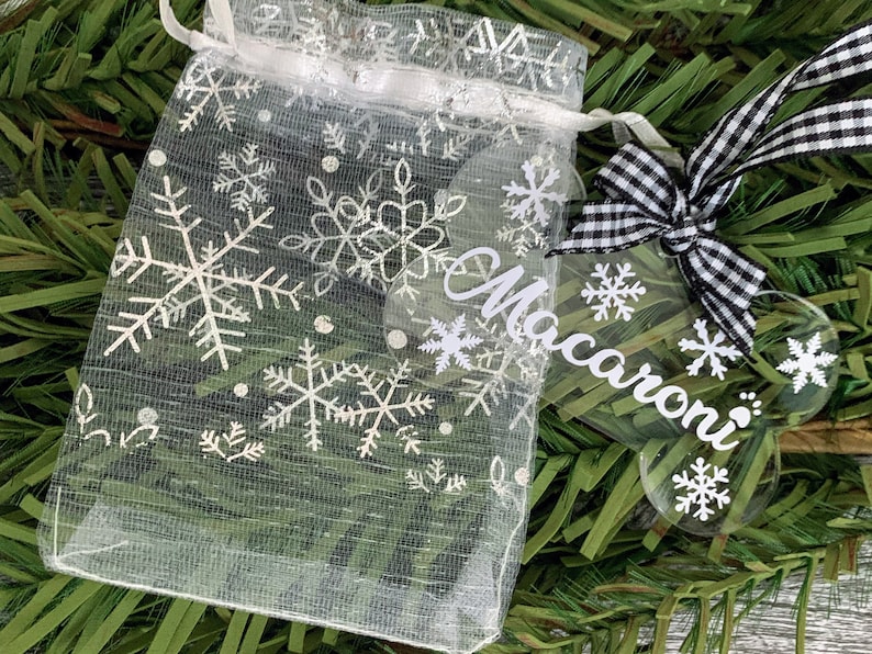 May include: A clear acrylic bone-shaped Christmas bauble with the name "Macaroni" written on it in white. The bauble is decorated with white snowflakes and is tied with a black and white checked ribbon.