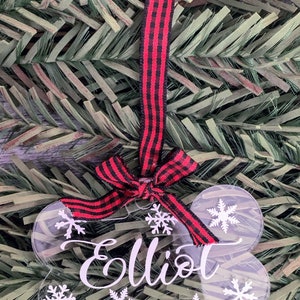 May include: A clear acrylic dog bone ornament with the name "Elliot" written in white. The ornament is decorated with white snowflakes and has a red and black checked ribbon attached.