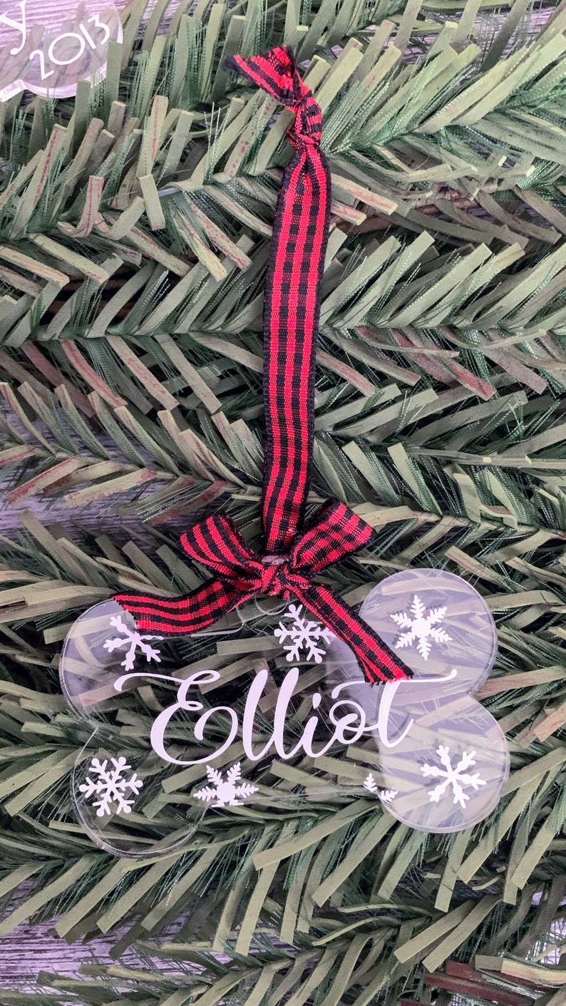 May include: A clear acrylic dog bone ornament with the name "Elliot" written in white. The ornament is decorated with white snowflakes and has a red and black checked ribbon attached.