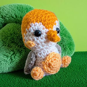May include: A handmade crochet penguin doll with orange, white, and gray yarn. The penguin has large black eyes and an orange beak and feet. The doll is sitting on a green surface with a green background.