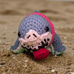 Puede incluir: Un juguete de tiburón tejido a mano en tonos azules, rosas y blancos, con una bola roja. El tiburón está sobre una playa arenosa con el océano al fondo. El juguete tiene ojos negros y una boca detallada.