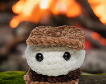 May include: A plush toy shaped like a s'more. The toy is made of brown, white, and brown yarn and has googly eyes. The toy is sitting on a wooden surface.