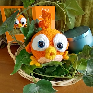 May include: Orange crocheted bird with white eyes and a yellow beak. The bird is sitting in a wicker basket with green leaves.