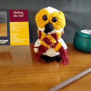 May include: A white crocheted owl with yellow eyes and a red and yellow scarf. The owl is sitting on a wooden surface with a wand and a teal jar in the background. The image includes a card with the text "Hedwig the Owl" and a list of materials needed to make the owl.