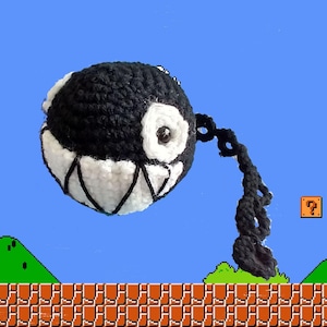May include: A black and white crocheted plush toy shaped like a character from the Super Mario Bros. video game series. The toy is hanging from a chain and is positioned above a brick wall and green hills.