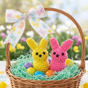 May include: A wicker basket filled with green paper grass, colorful Easter eggs, and two crocheted bunny figures. A yellow bunny and a pink bunny sit in the basket. A decorative bow with bunny and chick patterns adorns the basket.