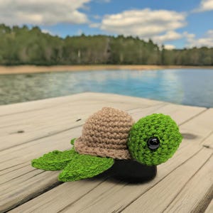 May include: A crocheted green and brown turtle with black eyes sits on a wooden dock. The turtle is facing the right side of the image. The background is a blurry image of a lake and trees.