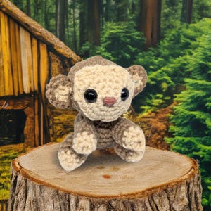 May include: A crocheted monkey doll in light brown and beige. The handmade toy has large black eyes and a small pink nose. The monkey is sitting on a tree stump in front of a blurred forest background.