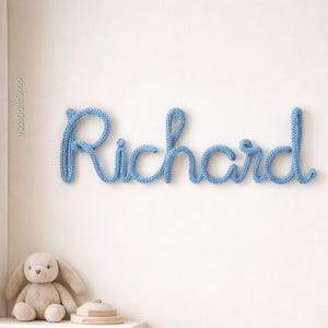 May include: A light blue, knitted wall hanging spelling the name "Richard" in cursive script. The name is mounted on a white wall. A stuffed bunny and wooden toy are on a shelf below.