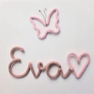 May include: A light pink knitted wire wall decoration spelling the name Eva in cursive script, accompanied by a separate light pink knitted wire heart and a light pink knitted wire butterfly outline with thin black wire antennae.