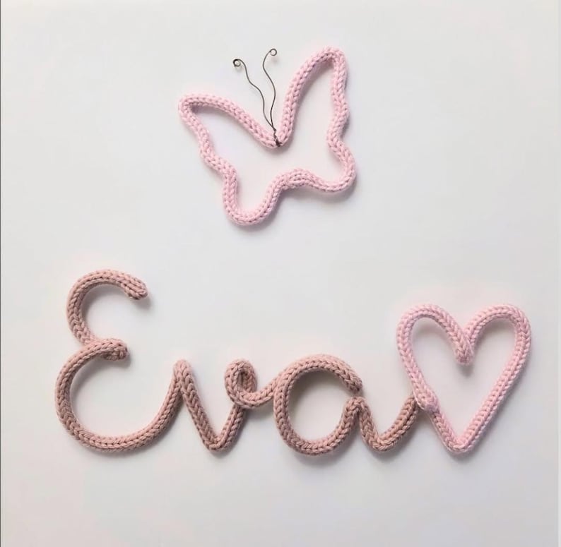 May include: A light pink knitted wire wall decoration spelling the name Eva in cursive script, accompanied by a separate light pink knitted wire heart and a light pink knitted wire butterfly outline with thin black wire antennae.
