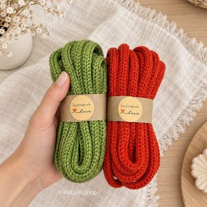 May include: Two bundles of thick, knitted cord in olive green and bright red. Each bundle is secured with a brown paper band featuring the text 'handmade with love' and a small red heart icon. The cords have a visible, textured knit pattern.