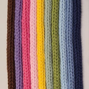 May include: A collection of nine vertical knitted cords arranged side by side in a spectrum of colors including brown, purple, magenta, light pink, yellow, sage green, olive green, light blue, and dark navy blue with a visible braided texture.