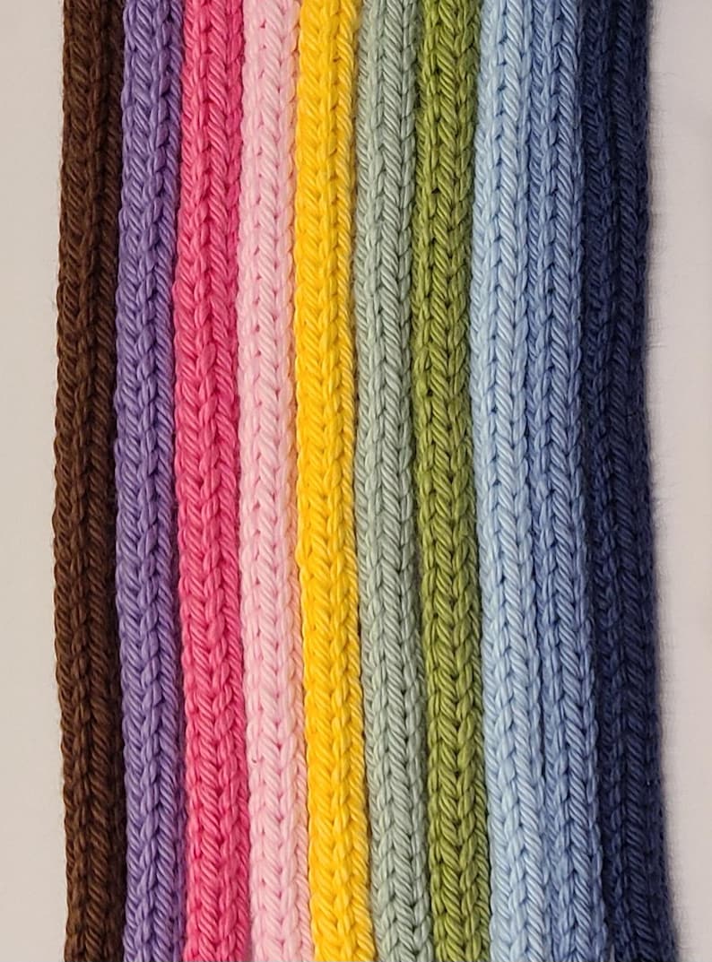 May include: A collection of nine vertical knitted cords arranged side by side in a spectrum of colors including brown, purple, magenta, light pink, yellow, sage green, olive green, light blue, and dark navy blue with a visible braided texture.
